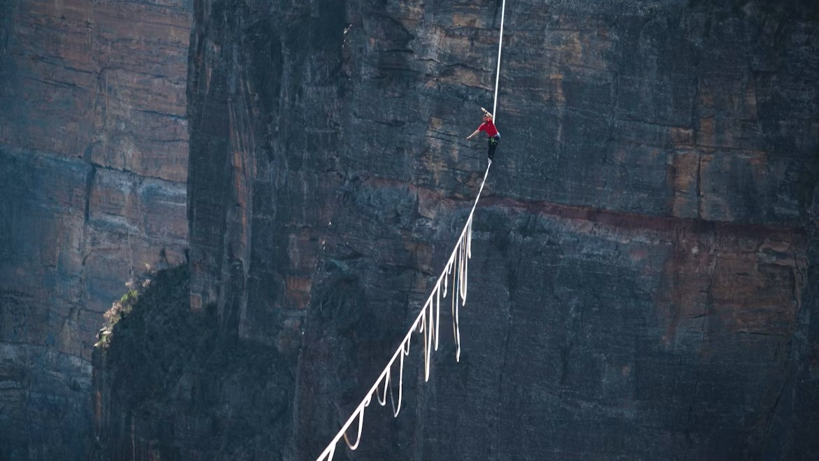A lone figure in a red shirt balances on a slackline strung high across a vast dark rockface, with the line and its trailing slack dropping away into the gorge far below.